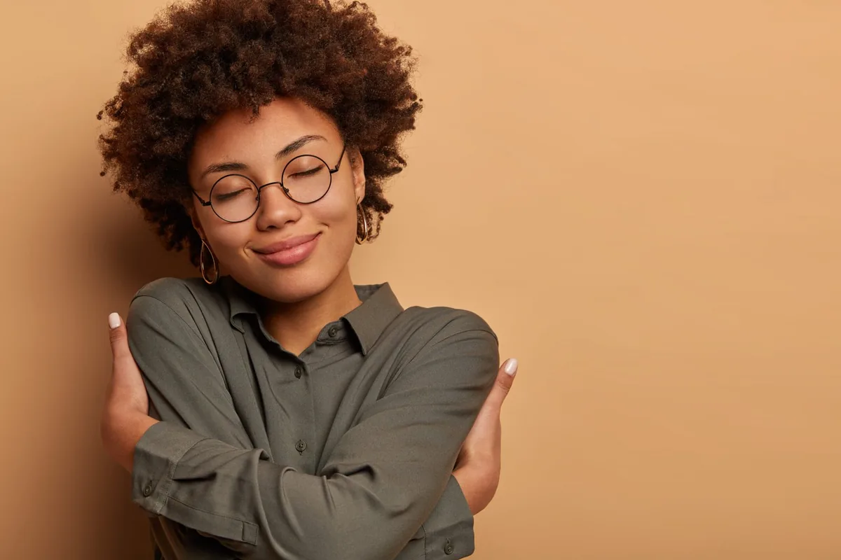 Mulher sorridente, de olhos fechados, abraçando a si mesma