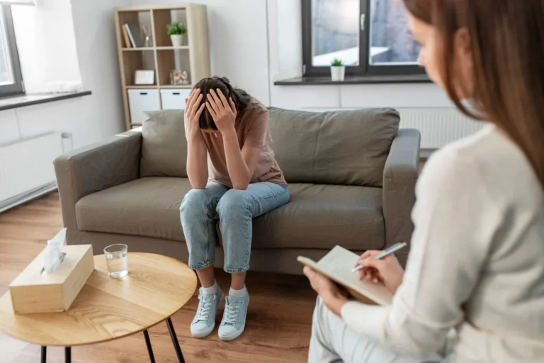 Paciente com as mãos na cabeça, sentada no sofá, conversando com uma psicóloga que faz anotações